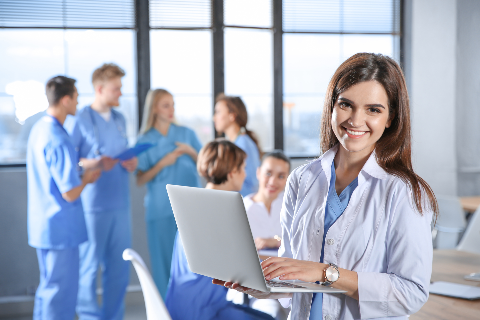 Medical student holding laptop, smiling at camera, group of nursing students in the background