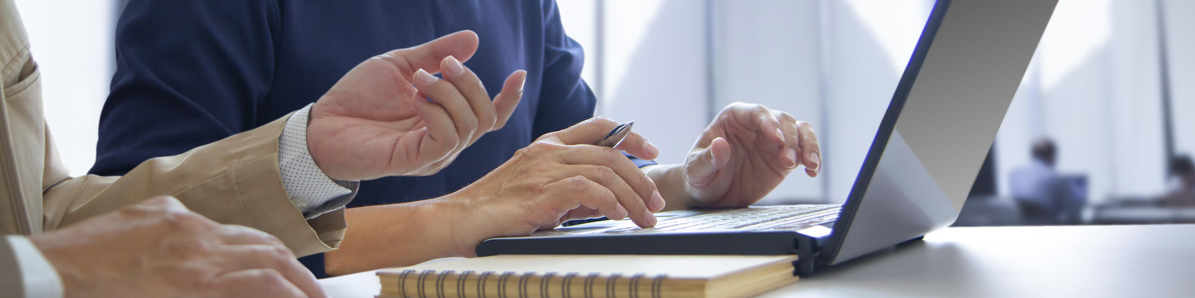 two people sitting side by side talking, one working on a laptop with a pen in hand and a pad of paper next to them.