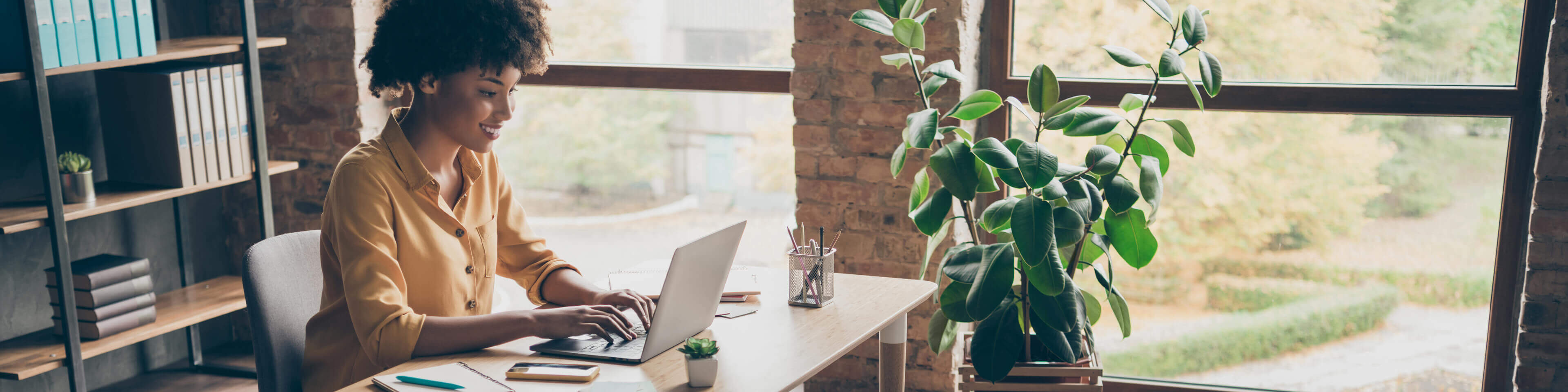 woman with a yellow button up shirt on sitting at a desk in a relaxed home office typing.