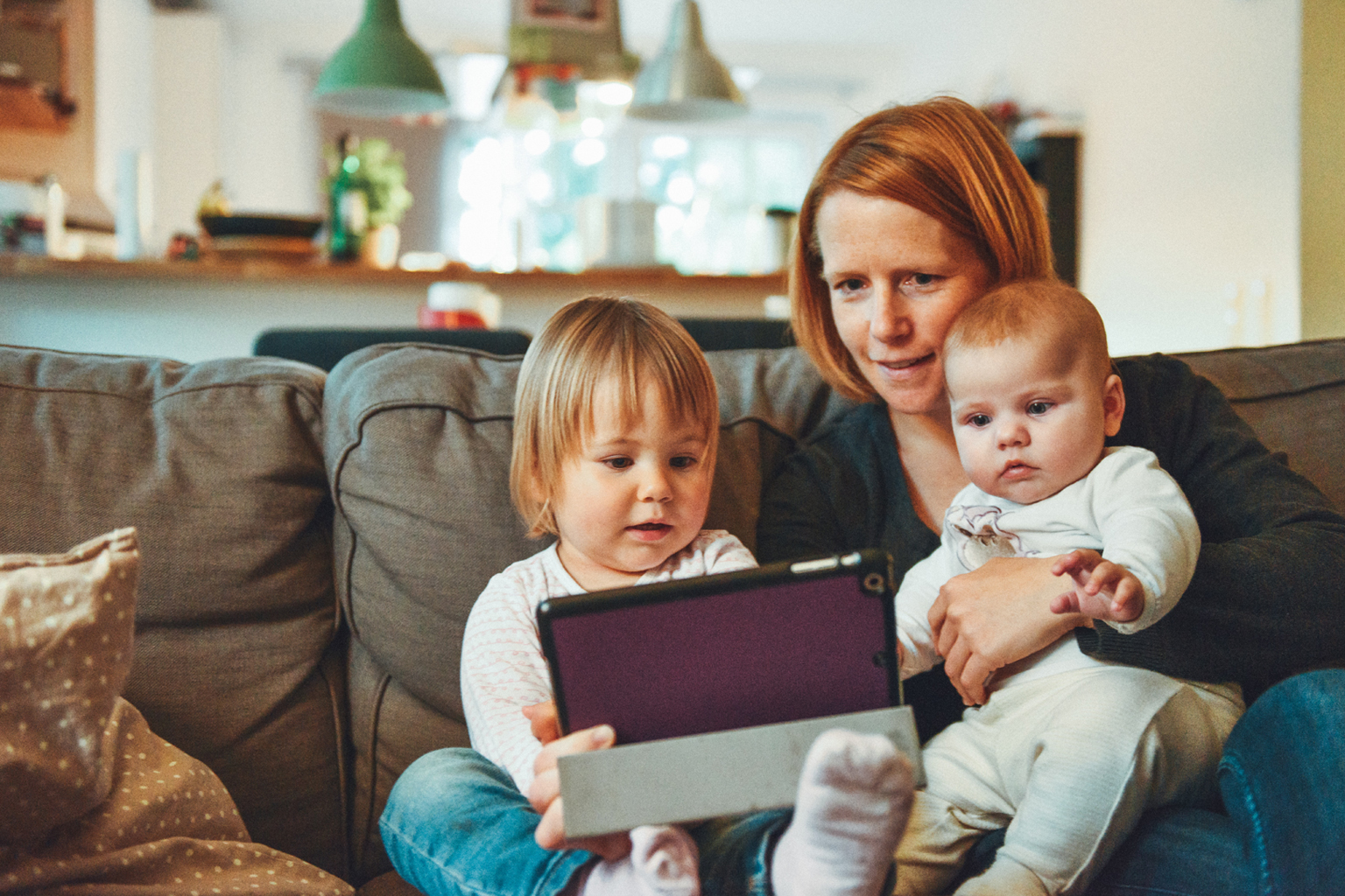 Woman sitting on couch with two young children, all watching tablet