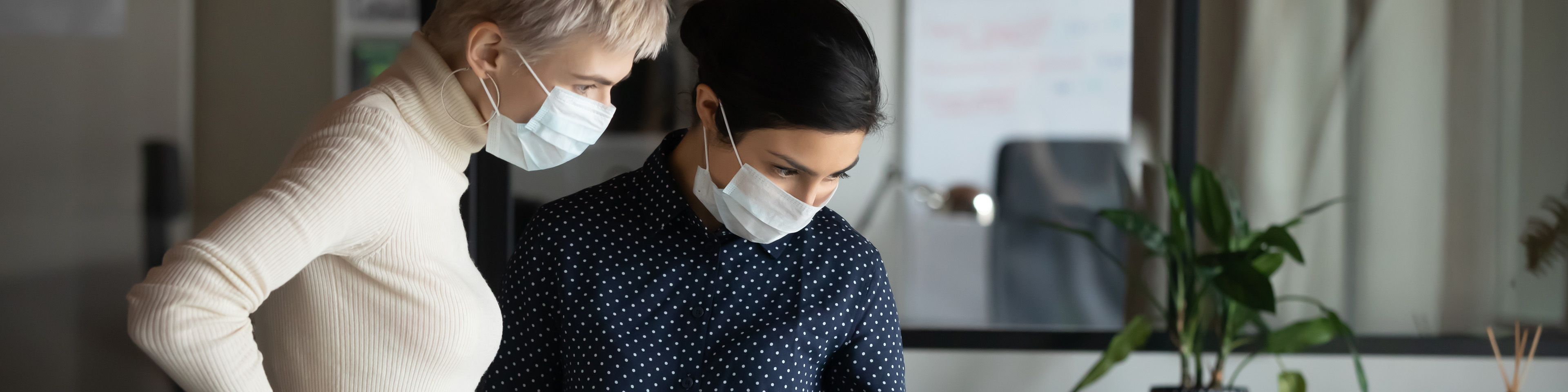 Focused two young multiracial business women in medical facial protective masks standing near table, looking at laptop screen, discussing project details