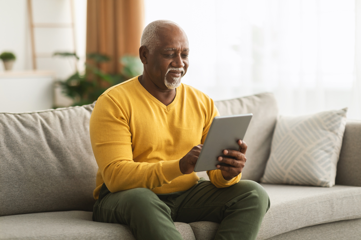 A pleasant older male sitting on a couch looking at a tablet.