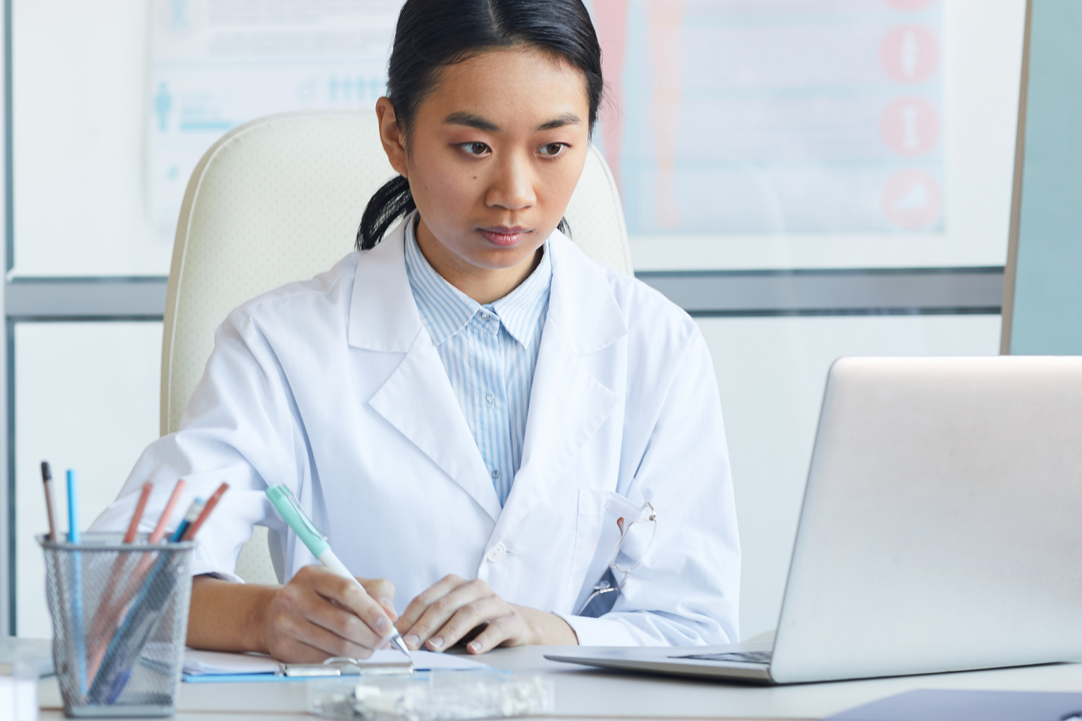 Portrait of female Asian doctor using laptop while sitting at workplace in medical office interior