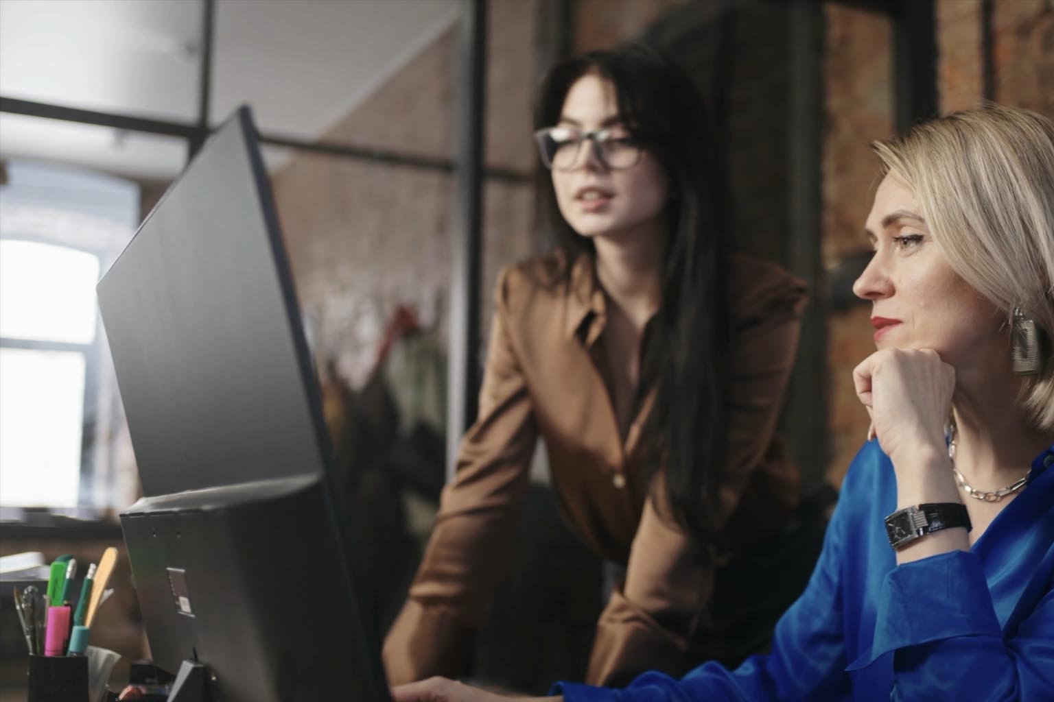 Two women, one seated and one standing, looking at a computer in a professional business setting