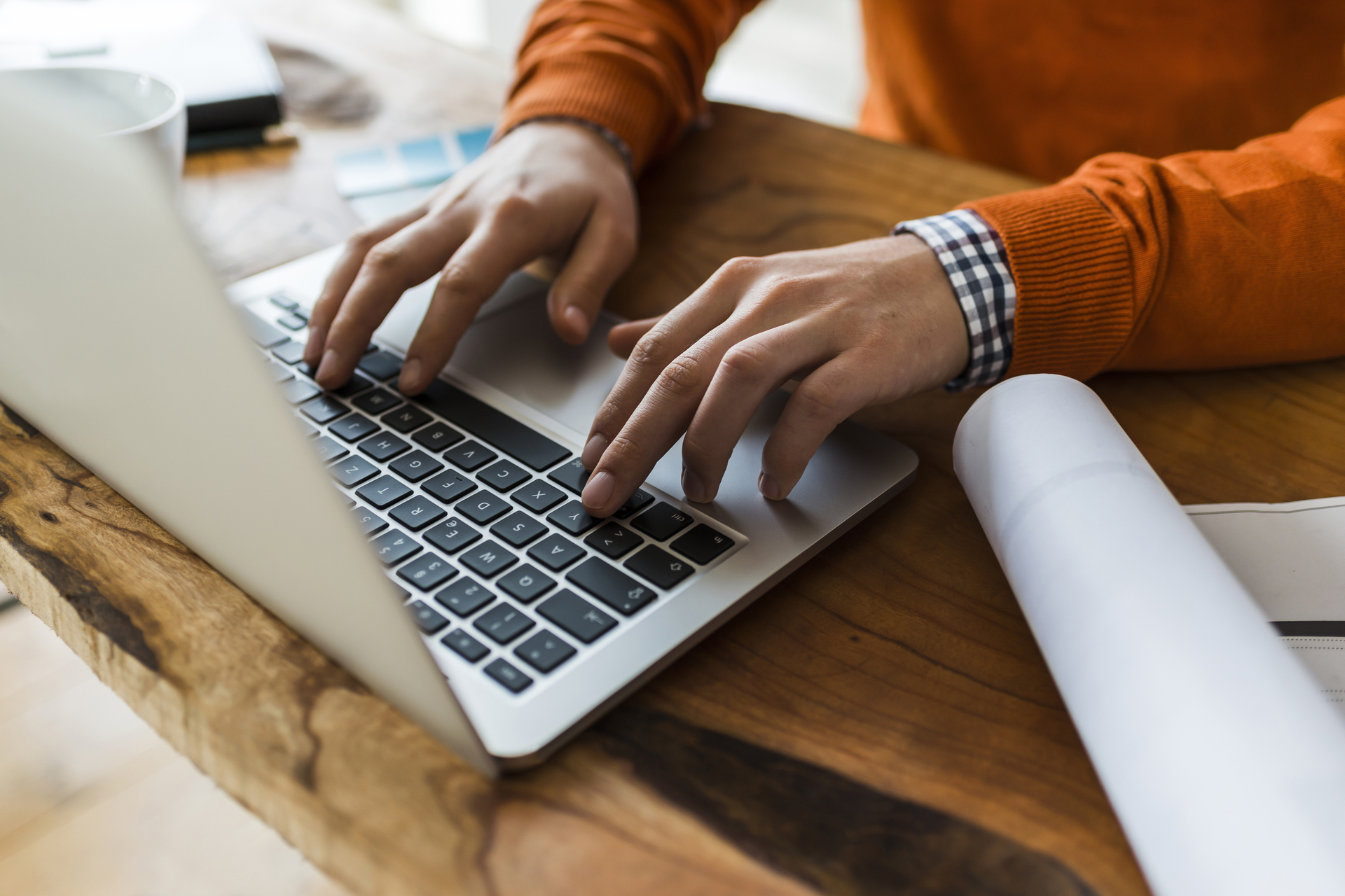 Man with orange sweatshirt, and flannel sitting at a wood table, typing on macbook with architectural plans on desk, Q3 2021, TAA NA US - Preparer