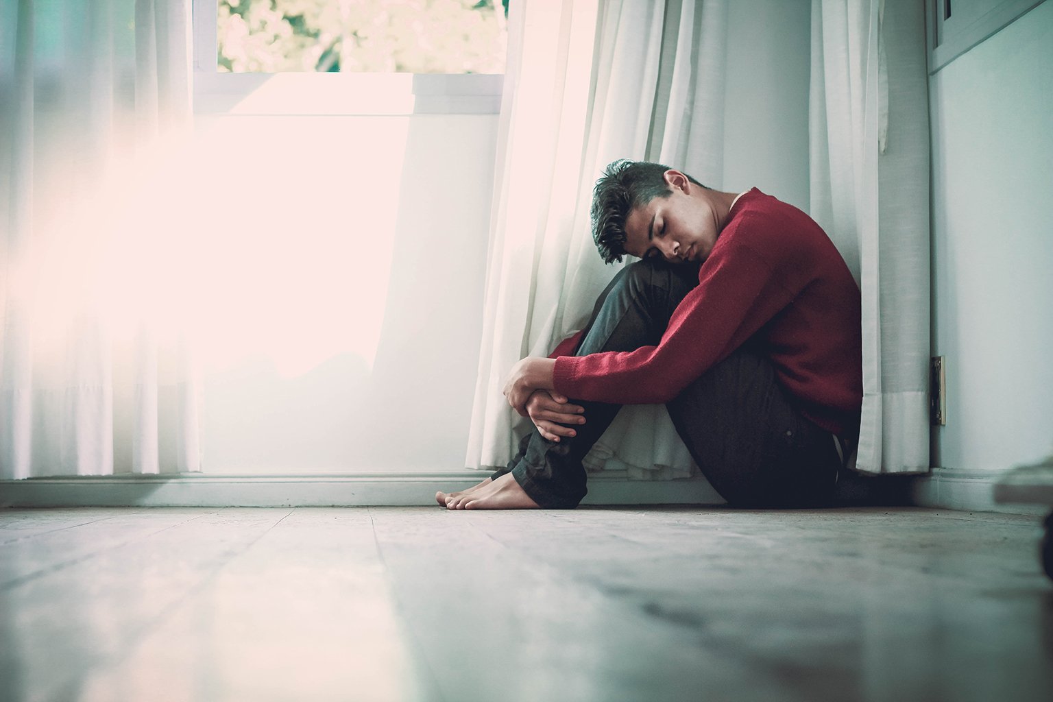 Young melancholy man sitting in corner, hugging his legs