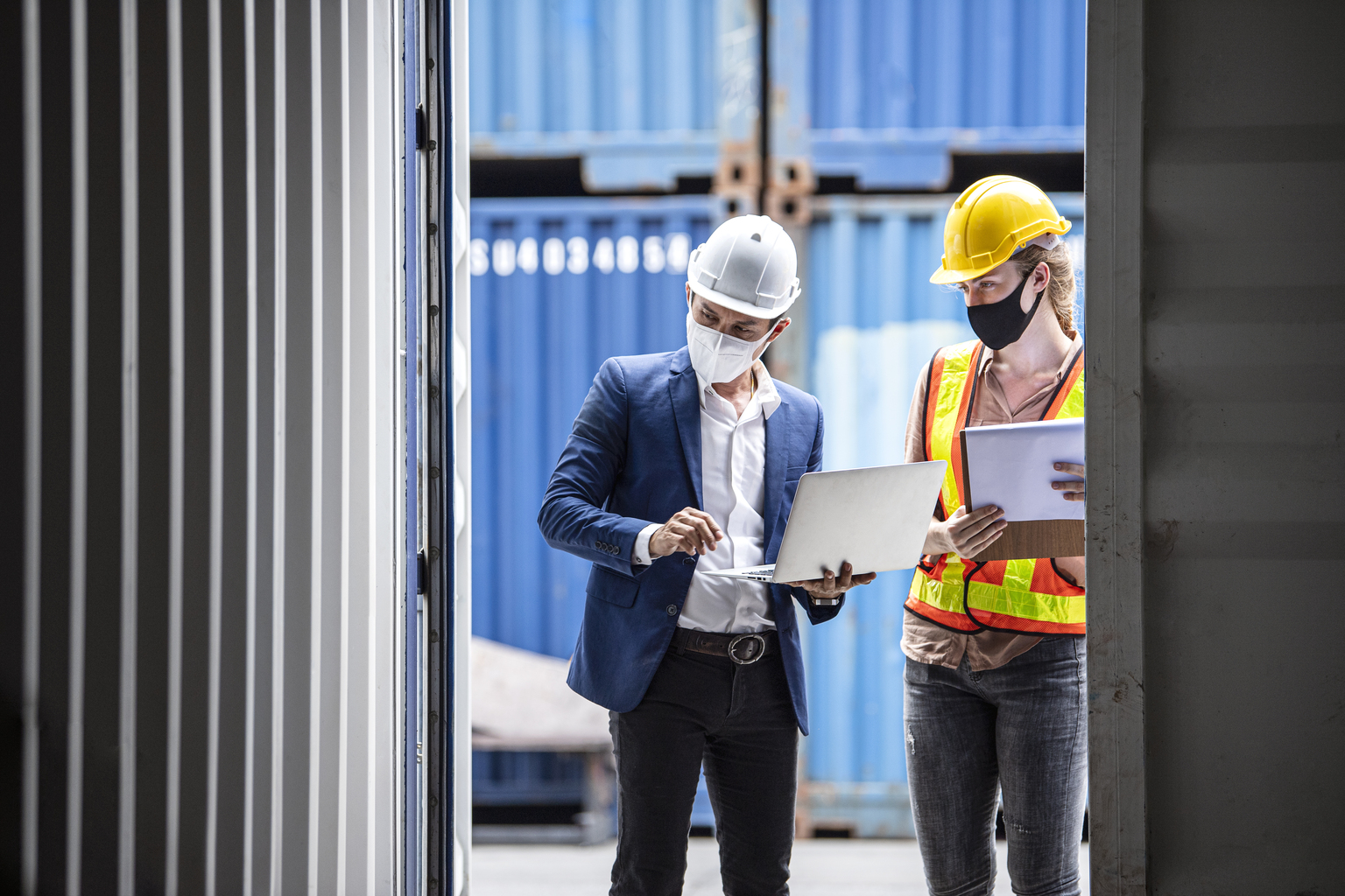 Business man and woman dock worker wearing safety protective clothing and face mask while working front of cargo container door open.