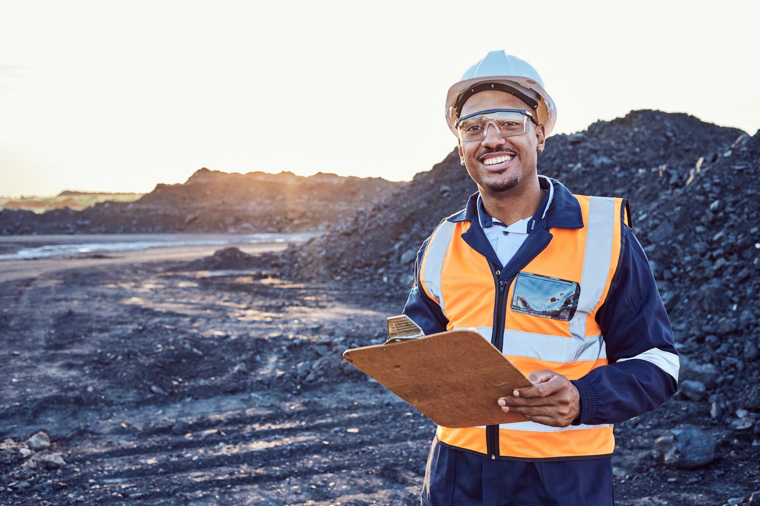 A young African mine worker wearing protective wear is looking at the camera while holding a clipboard with coal mine in the background