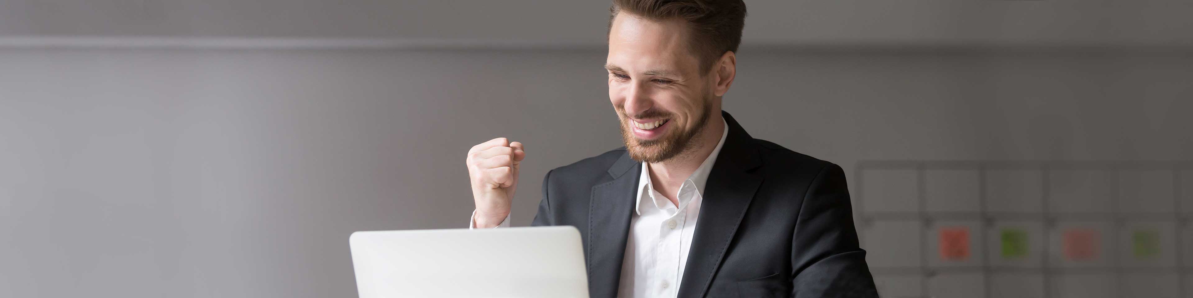man wearing grey suit jacket and white button up shirt with unbuttoned collar smiling and shaking his fist in excitement facing a laptop