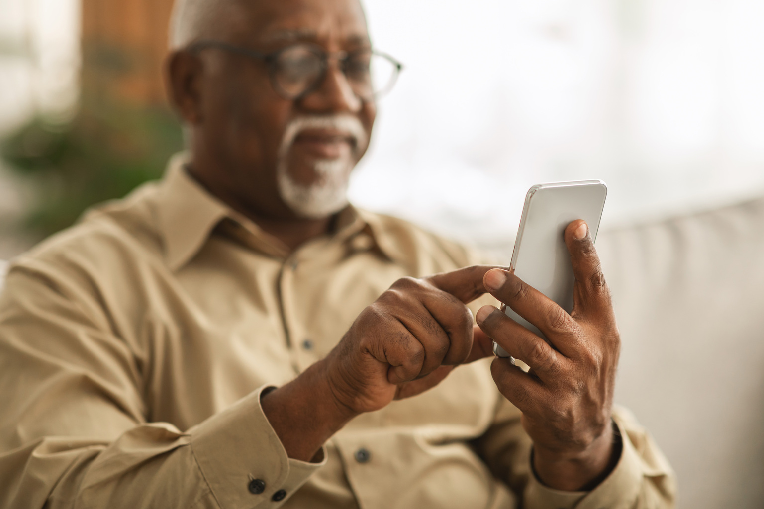 Senior African Man Using Smartphone Texting Sitting On Sofa Indoor