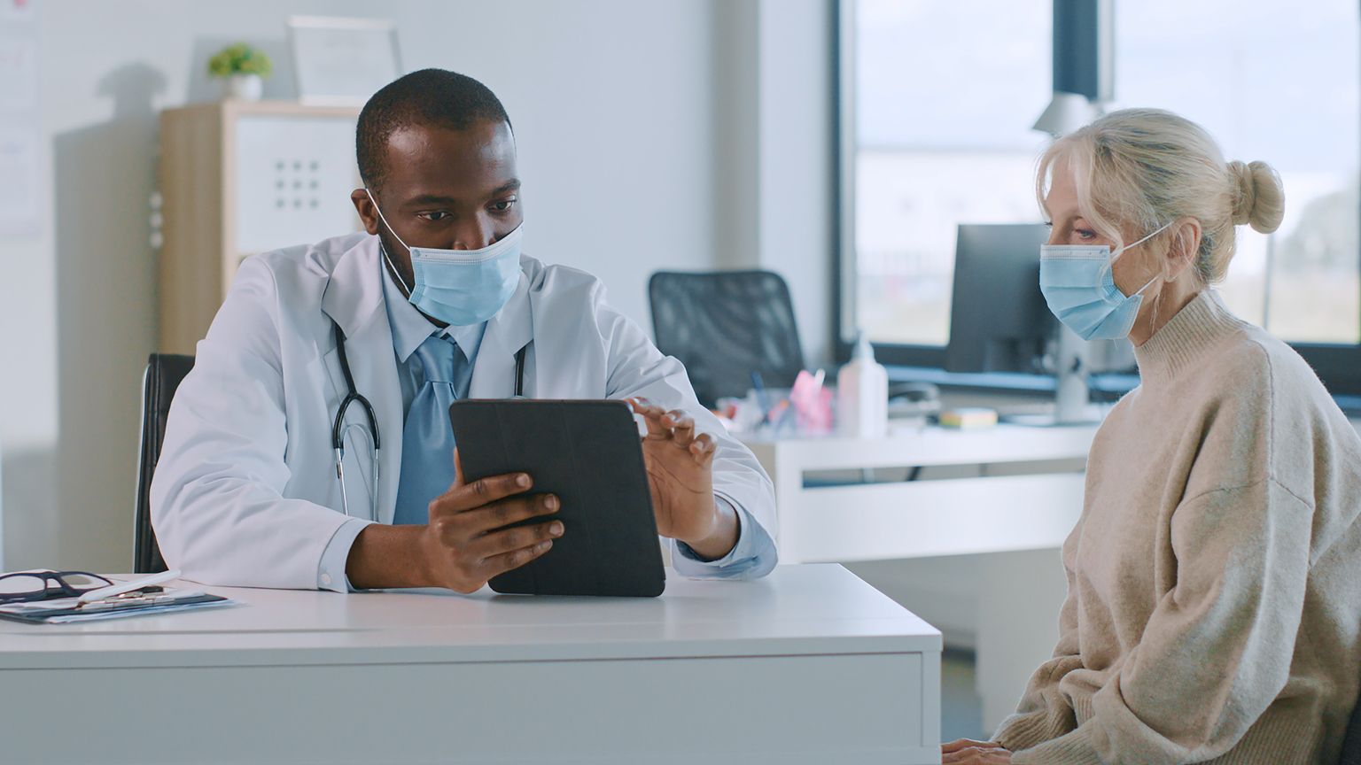 African American Doctor in Protective Mask is Reading Medical History of Senior Female Patient During Consultation in a Health Clinic