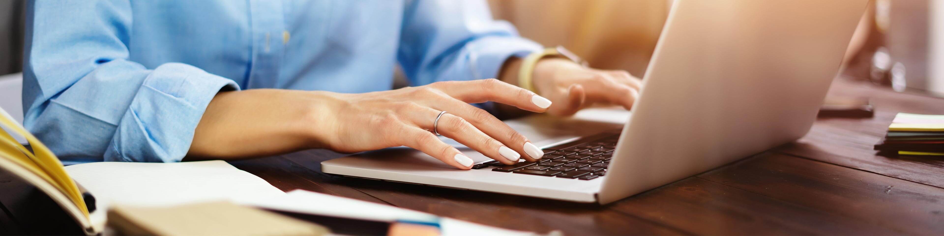 Woman in a blue button-up shirt typing on a laptop with paperwork next to her